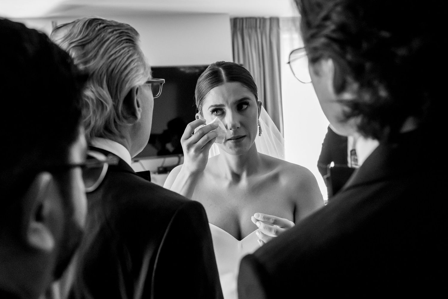 Black and white photo of a bride wiping tears while being comforted by loved ones during wedding preparations.