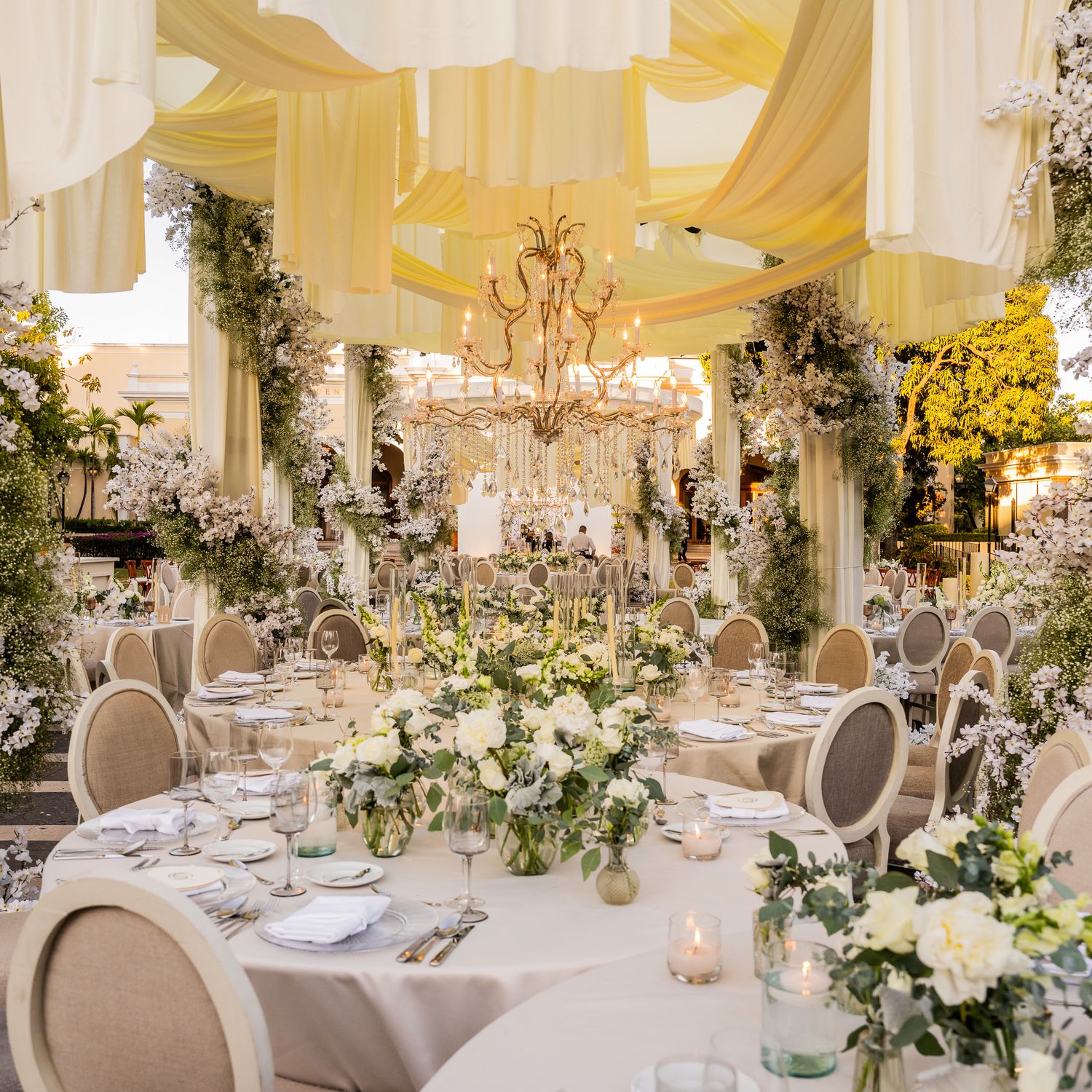 Elegant wedding reception hall with white draping, chandeliers and floral centerpieces on round tables.