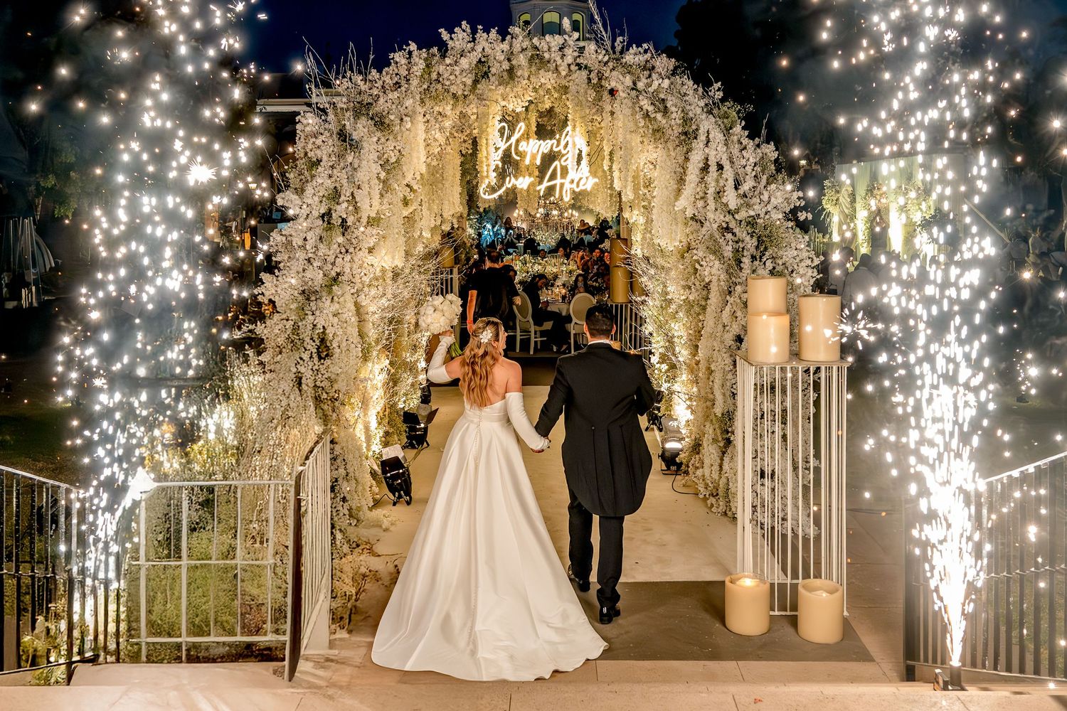 Bride and groom walk under a stunning white floral arch adorned with twinkling fairy lights at an elegant night wedding.