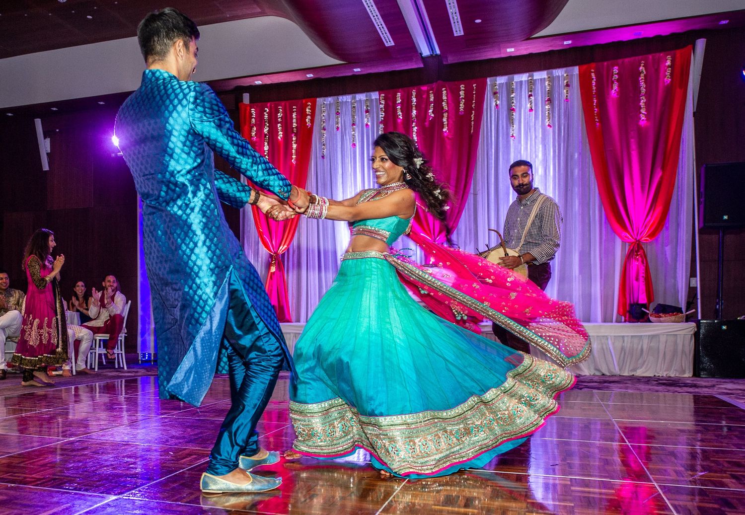 Couple dancing in vibrant teal and pink traditional Indian attire at a colorful wedding reception.