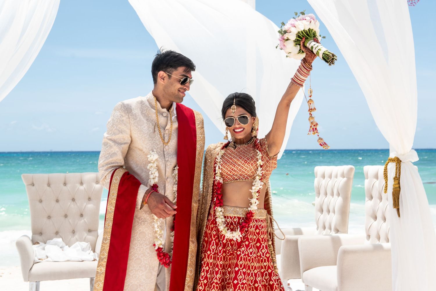 Indian bride in red lehenga raises bouquet joyfully beside groom in cream sherwani at beachfront wedding ceremony.