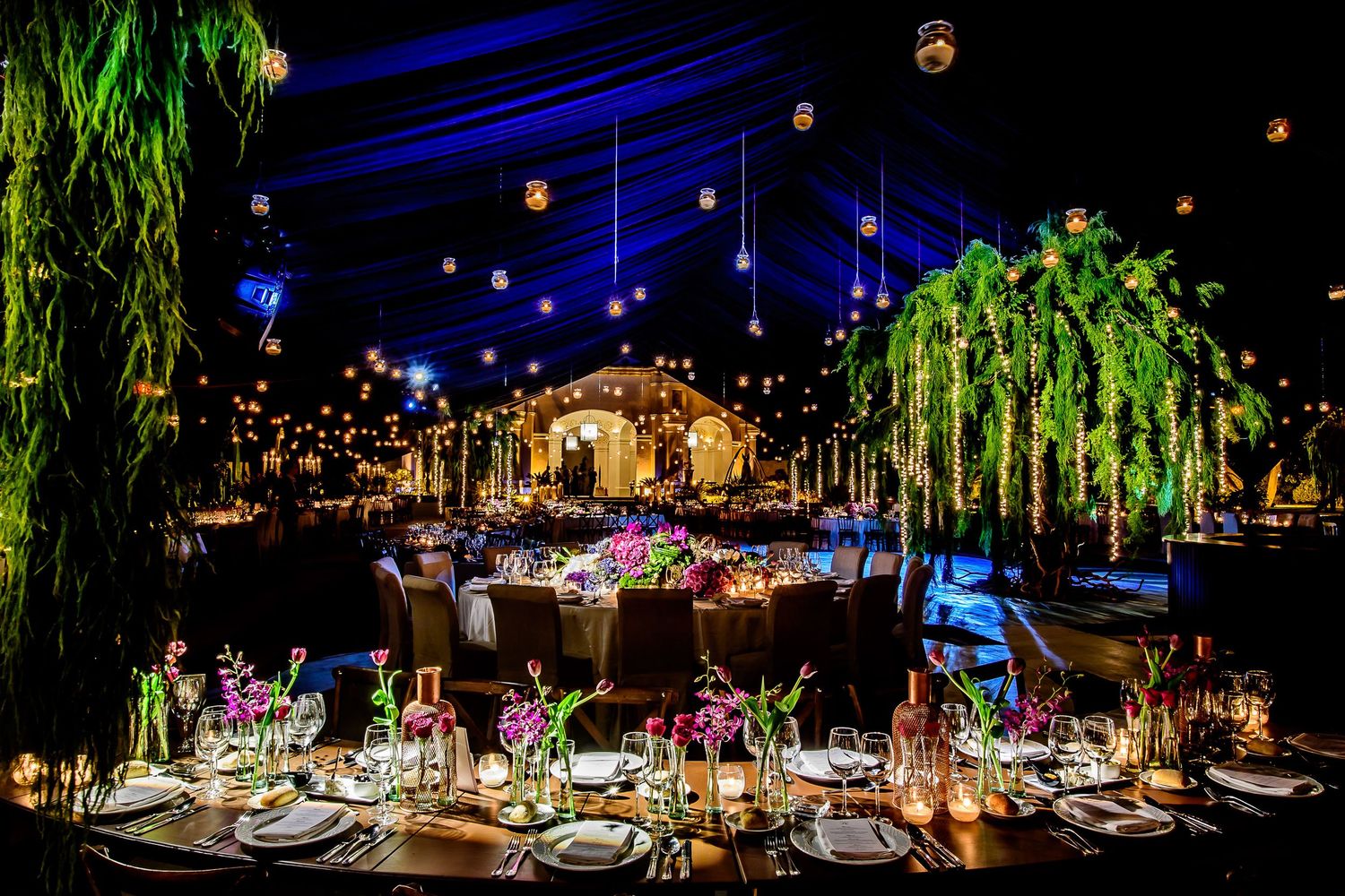 Elegant wedding reception under a dark tent with glowing lanterns, lush greenery centerpieces, and colorful floral table settings.