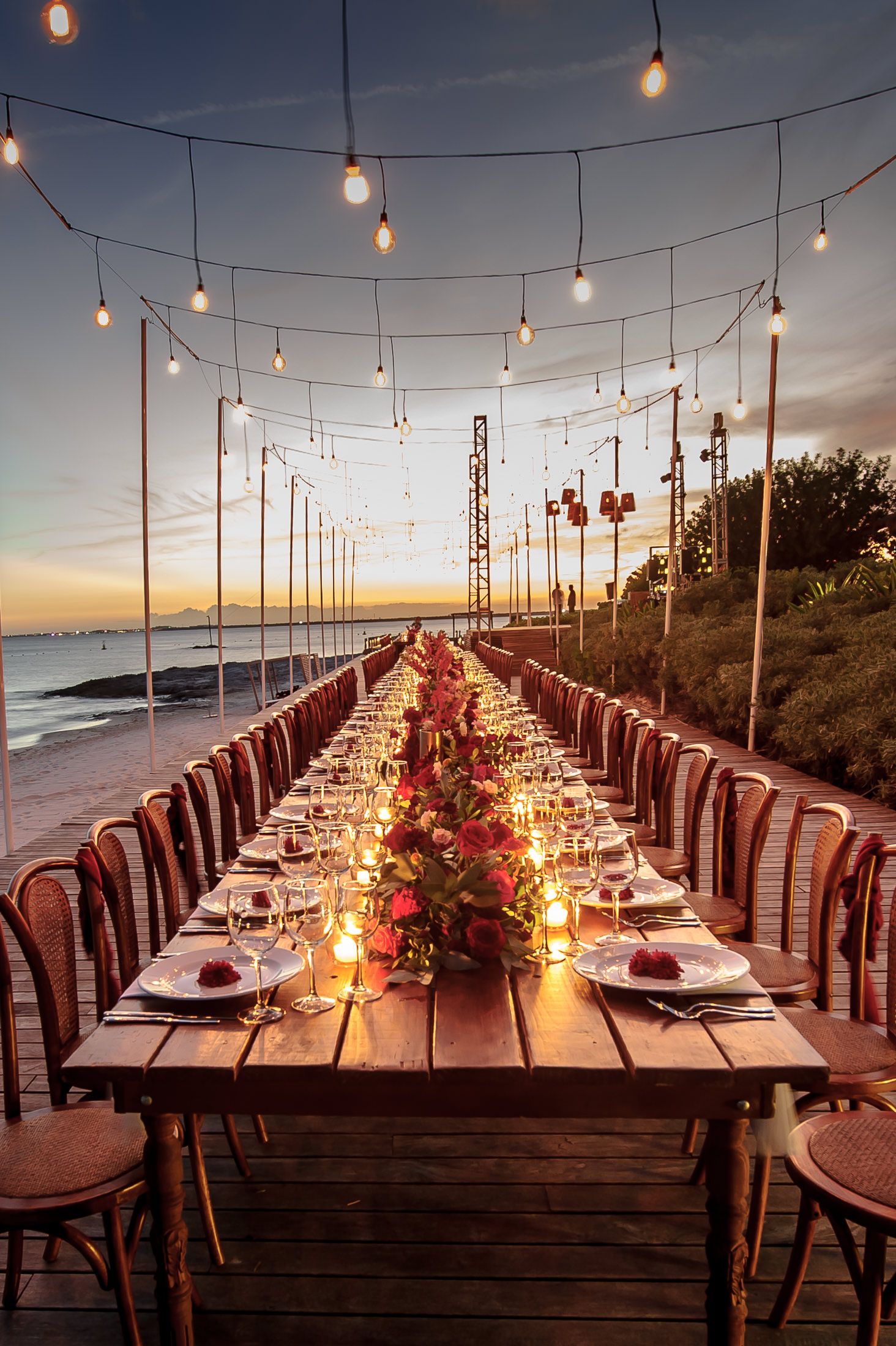 Romantic outdoor dining setup with string lights along a wooden deck overlooking the ocean at sunset.