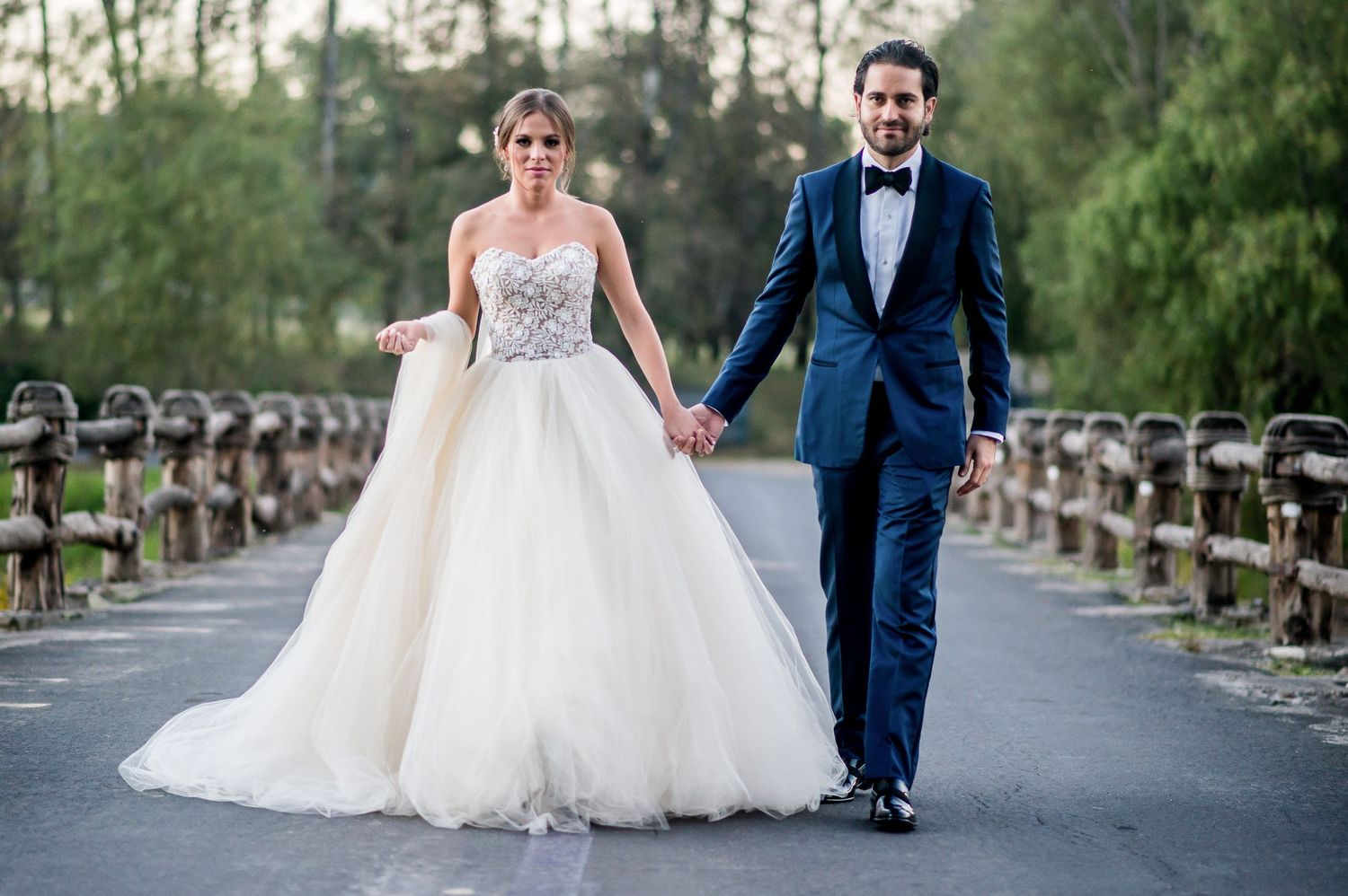 Elegant wedding couple walking along a tree-lined path in formal attire.