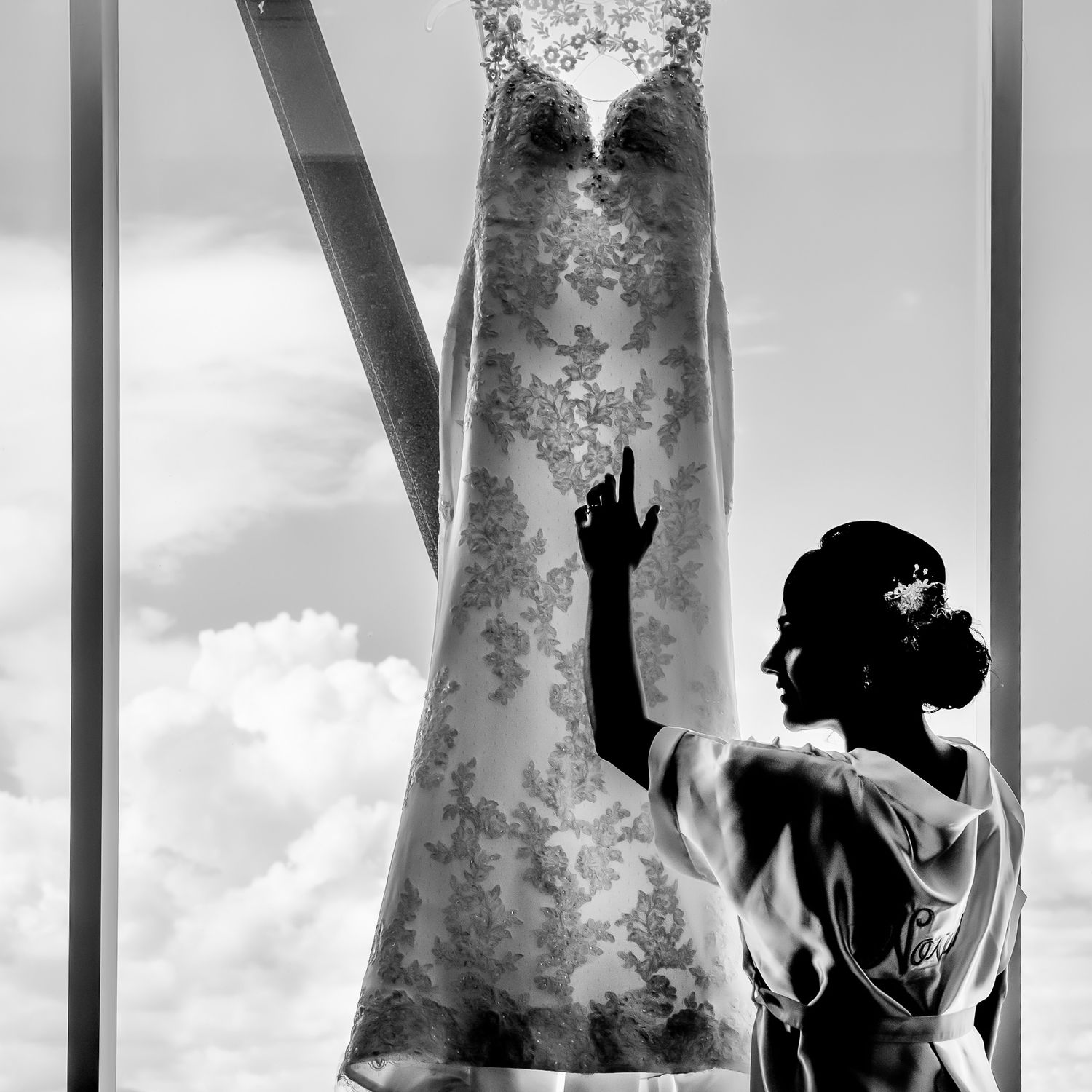 Wedding dress hanging in window with dramatic backlighting and clouds.