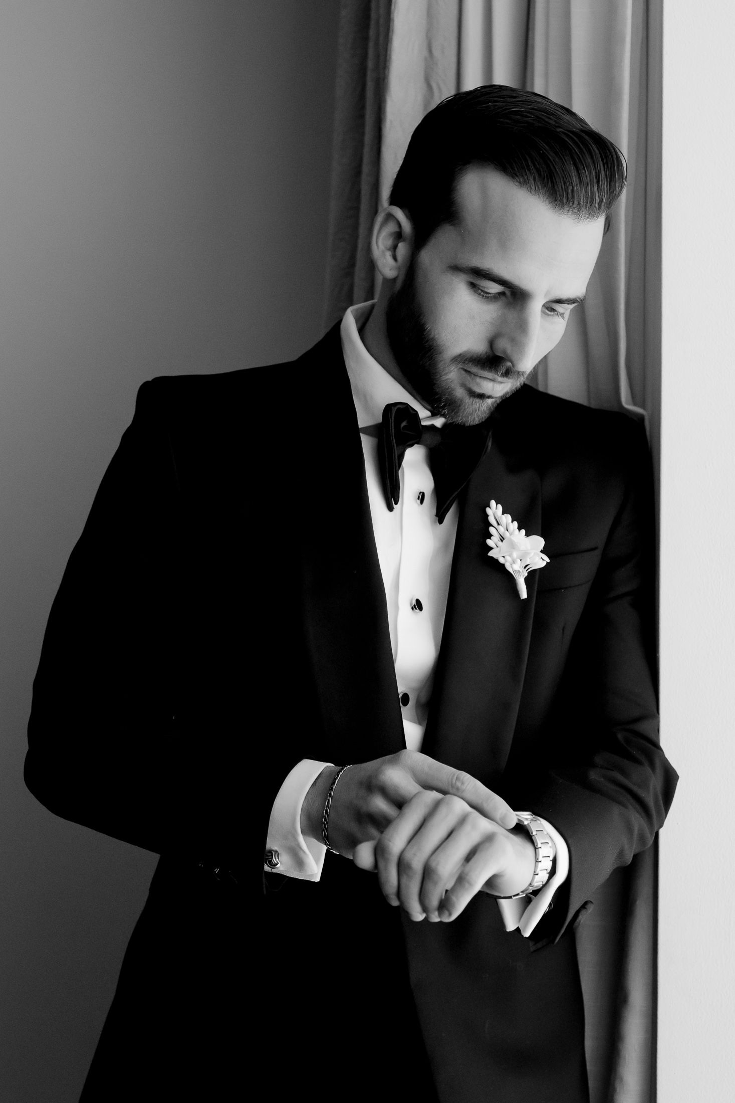 Elegant groom in black tuxedo adjusting cufflinks by window light.