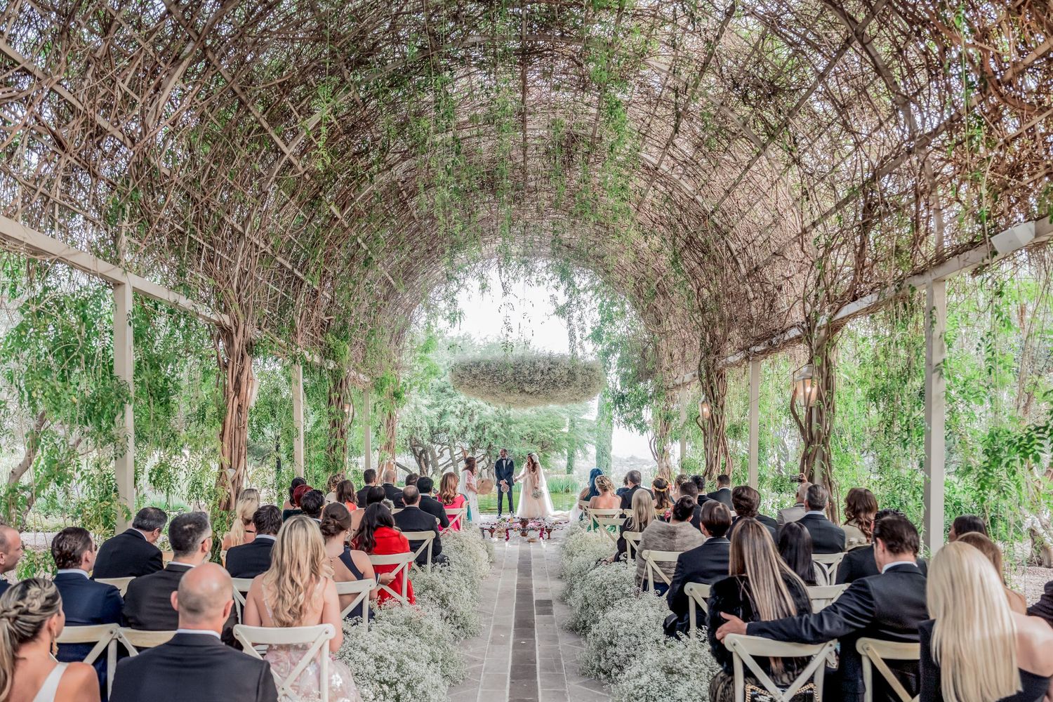 Wedding ceremony taking place under a lush garden arbor with guests seated on wooden chairs in an outdoor setting.