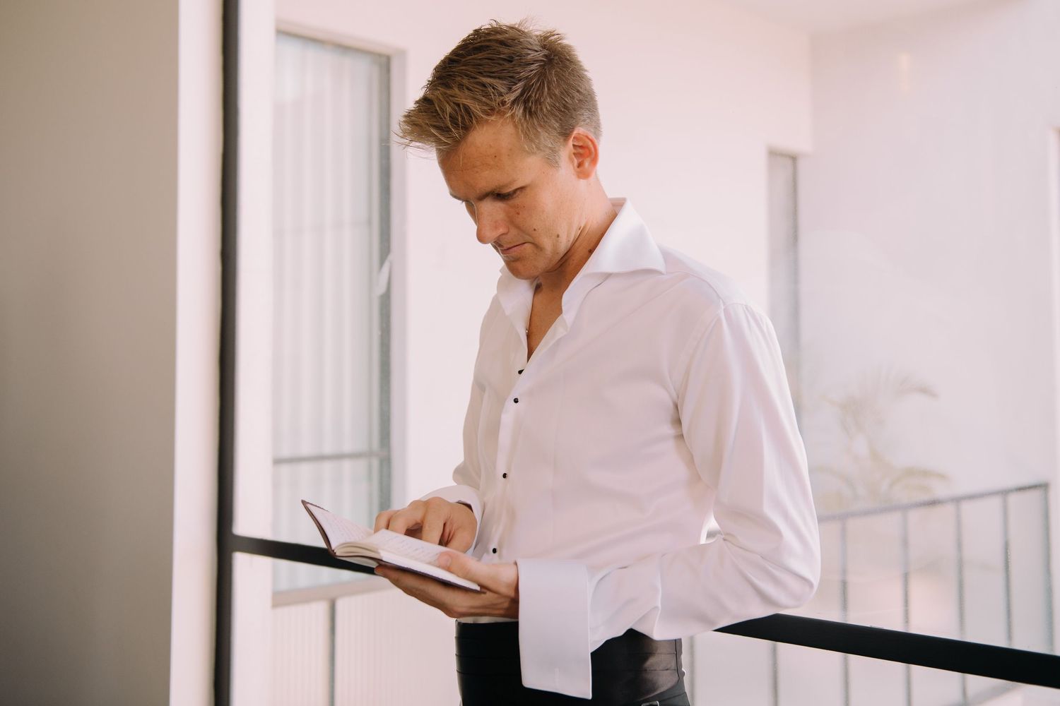 Professional man in white shirt reading a notebook while leaning on a railing in a bright modern office.