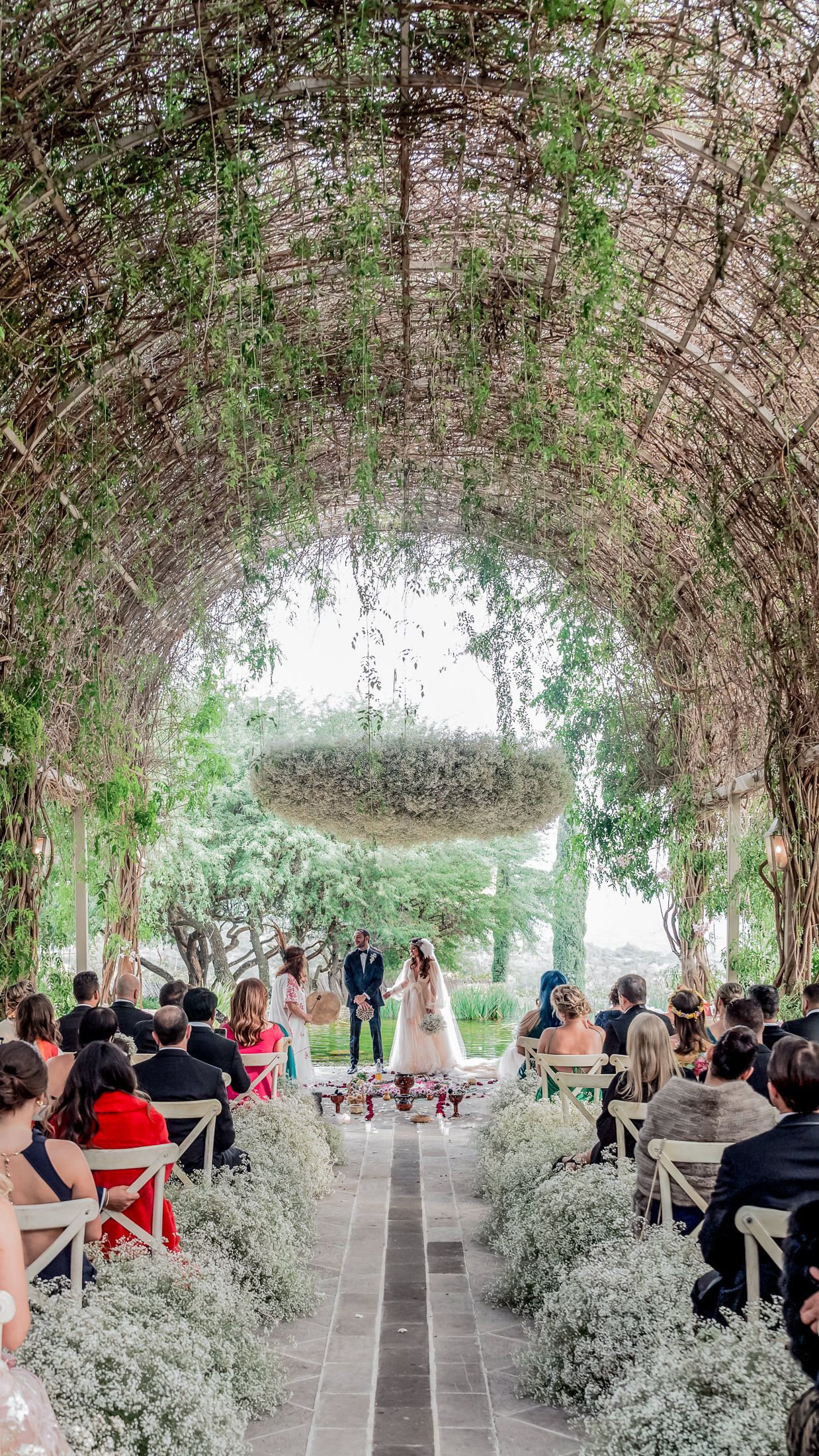 Elegant outdoor wedding ceremony under a lush green arched pergola with floral decorations and guests seated along the aisle.