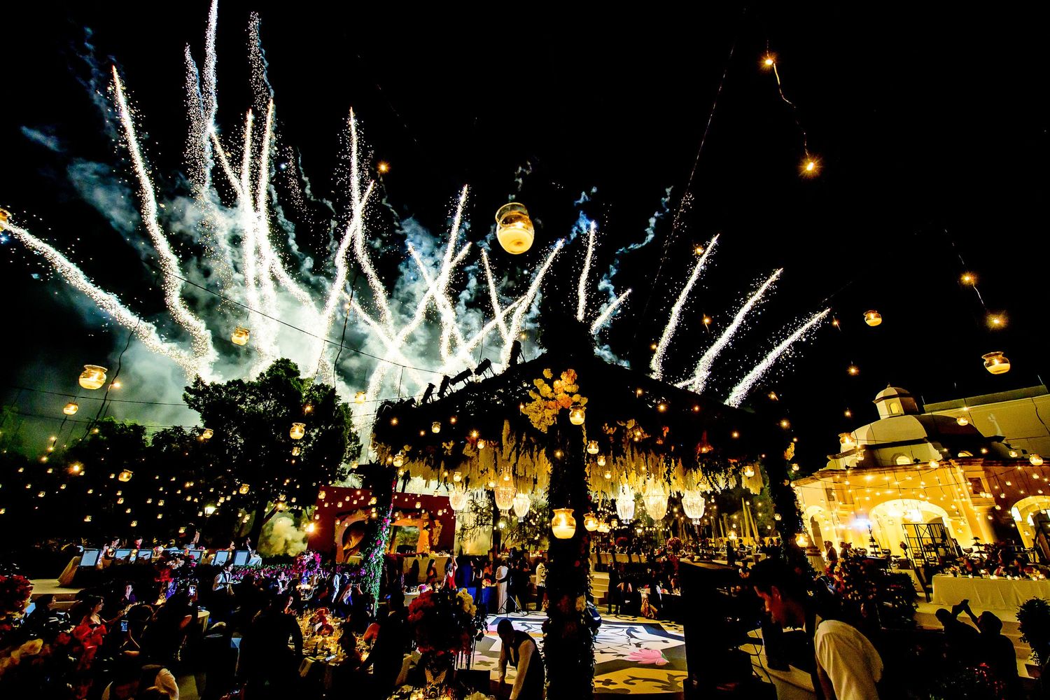 Colorful fireworks and floating lanterns light up the night sky above a festive crowd at an outdoor celebration.