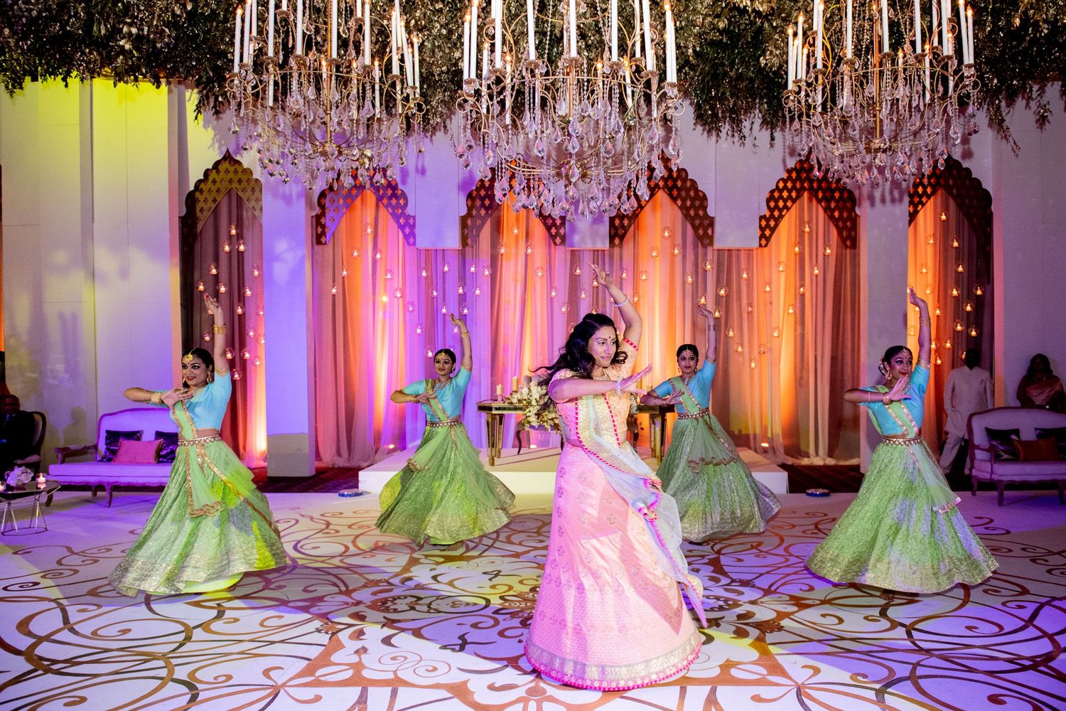 Bride in pink lehenga performs dance with green-clad bridesmaids under chandeliers at elegant Indian wedding reception.