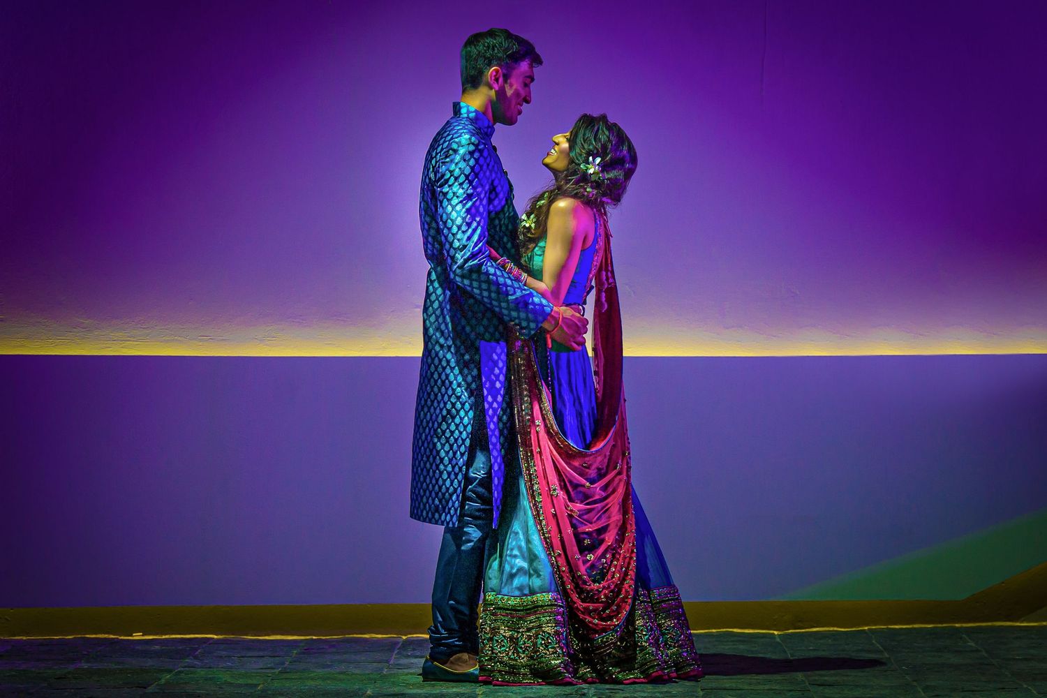 Couple in vibrant Indian wedding attire poses romantically against a purple and green gradient backdrop.