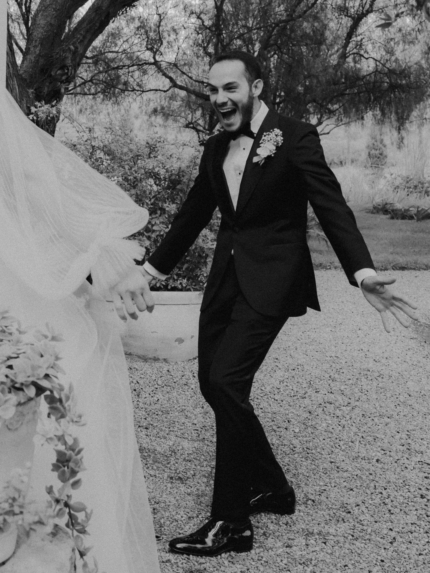 Groom in black tuxedo laughing with open arms during first look at outdoor wedding ceremony.