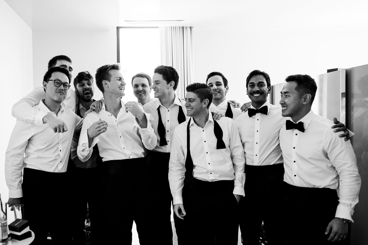 Group of groomsmen in white shirts and black bow ties laughing together before wedding ceremony.