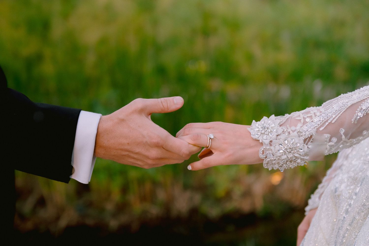 Bride and groom exchanging rings outdoors, close-up of hands with gold wedding band and lace bridal gown.