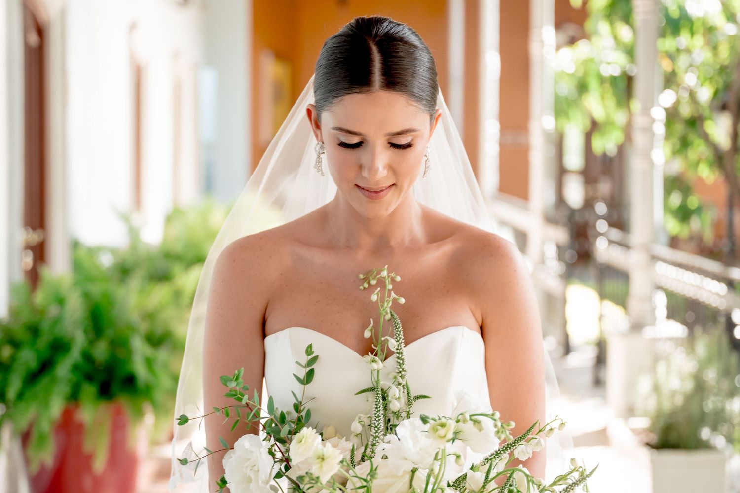 Elegant bride in strapless white gown holding white floral bouquet, wearing veil, looking down gracefully at her flowers.