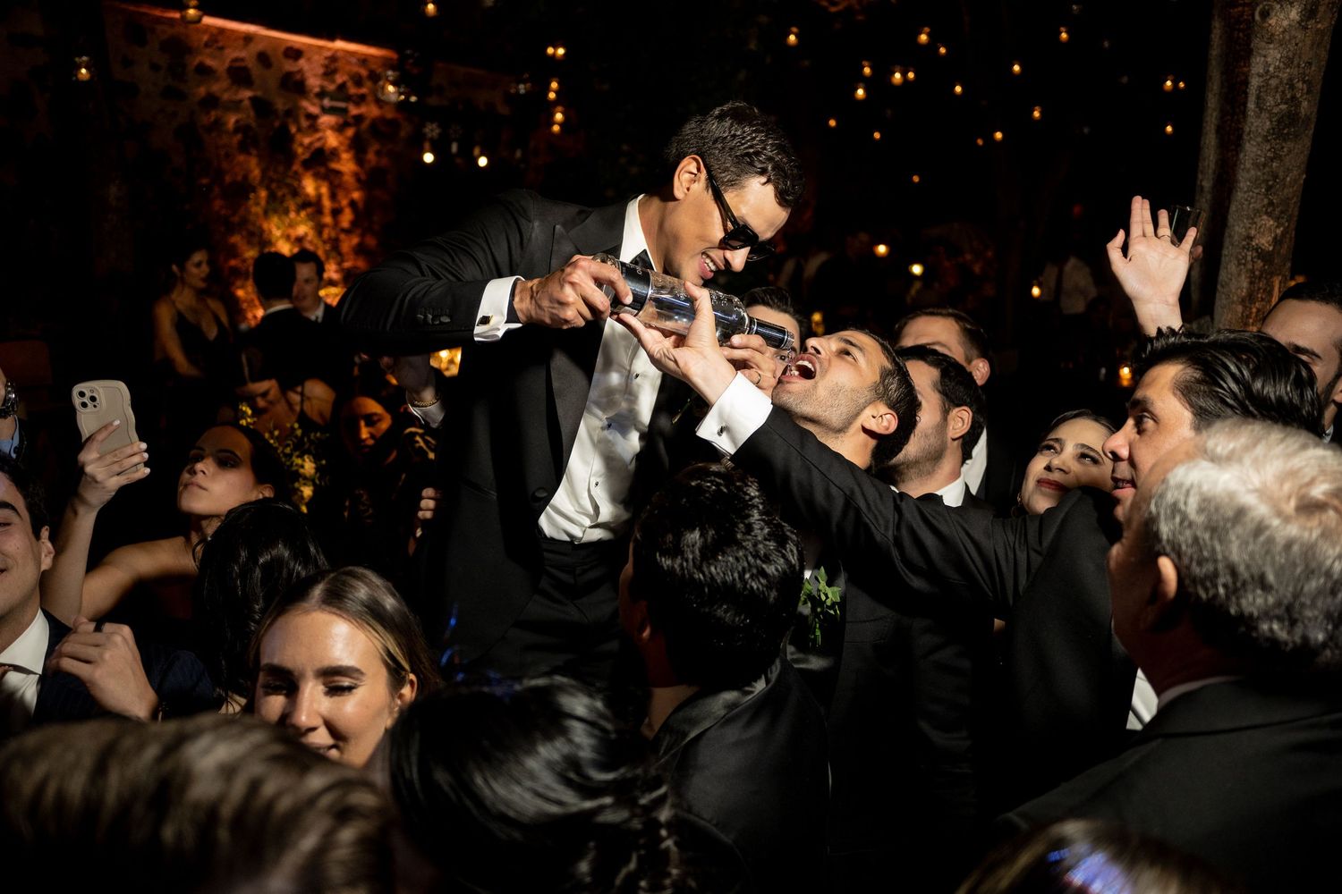Groom lifted by guests during lively wedding reception celebration at night.