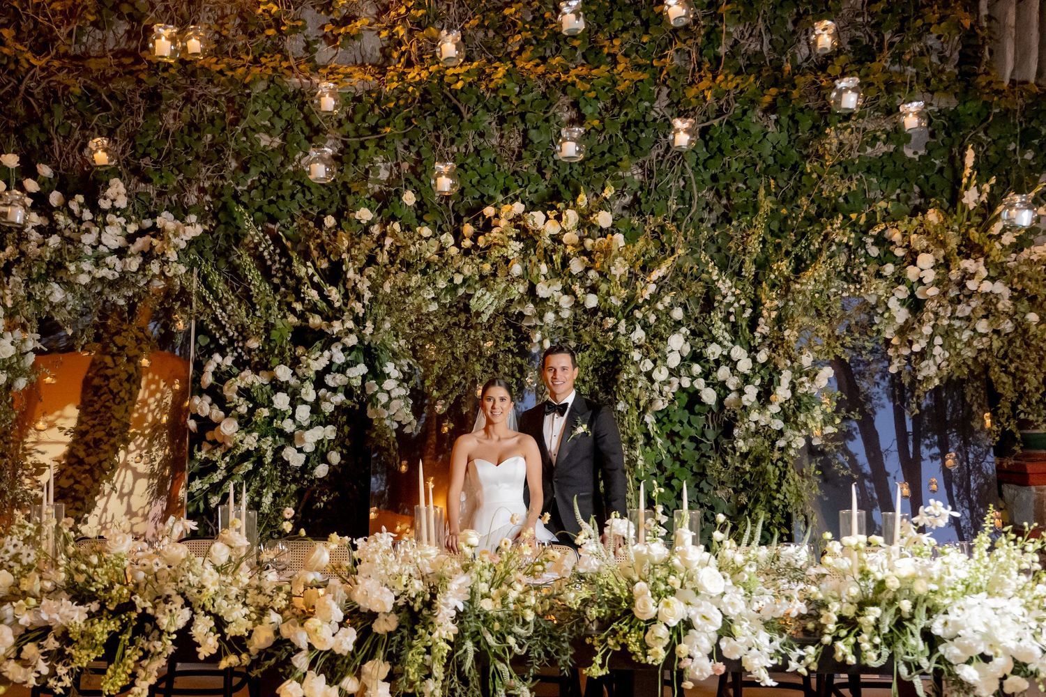 Bride and groom stand behind a sweetheart table adorned with white florals and candles beneath a lush greenery arch.