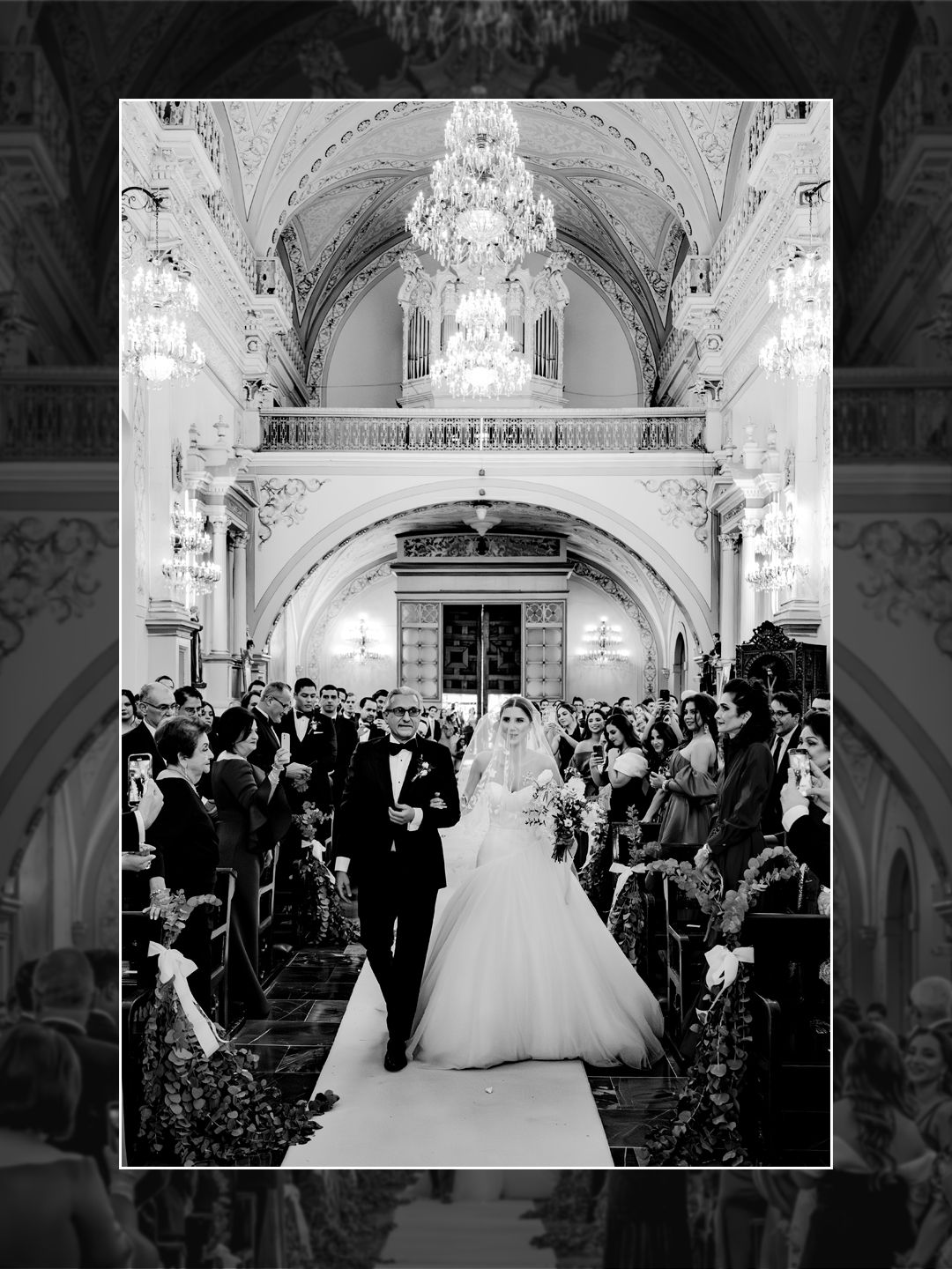 Black and white photo of a bride and groom walking down the aisle in an elegant grand ballroom with chandeliers.