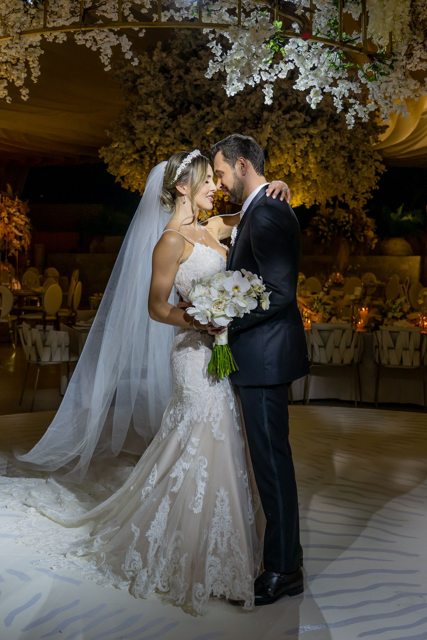 Elegant wedding couple shares intimate moment under floral decor on white dance floor.