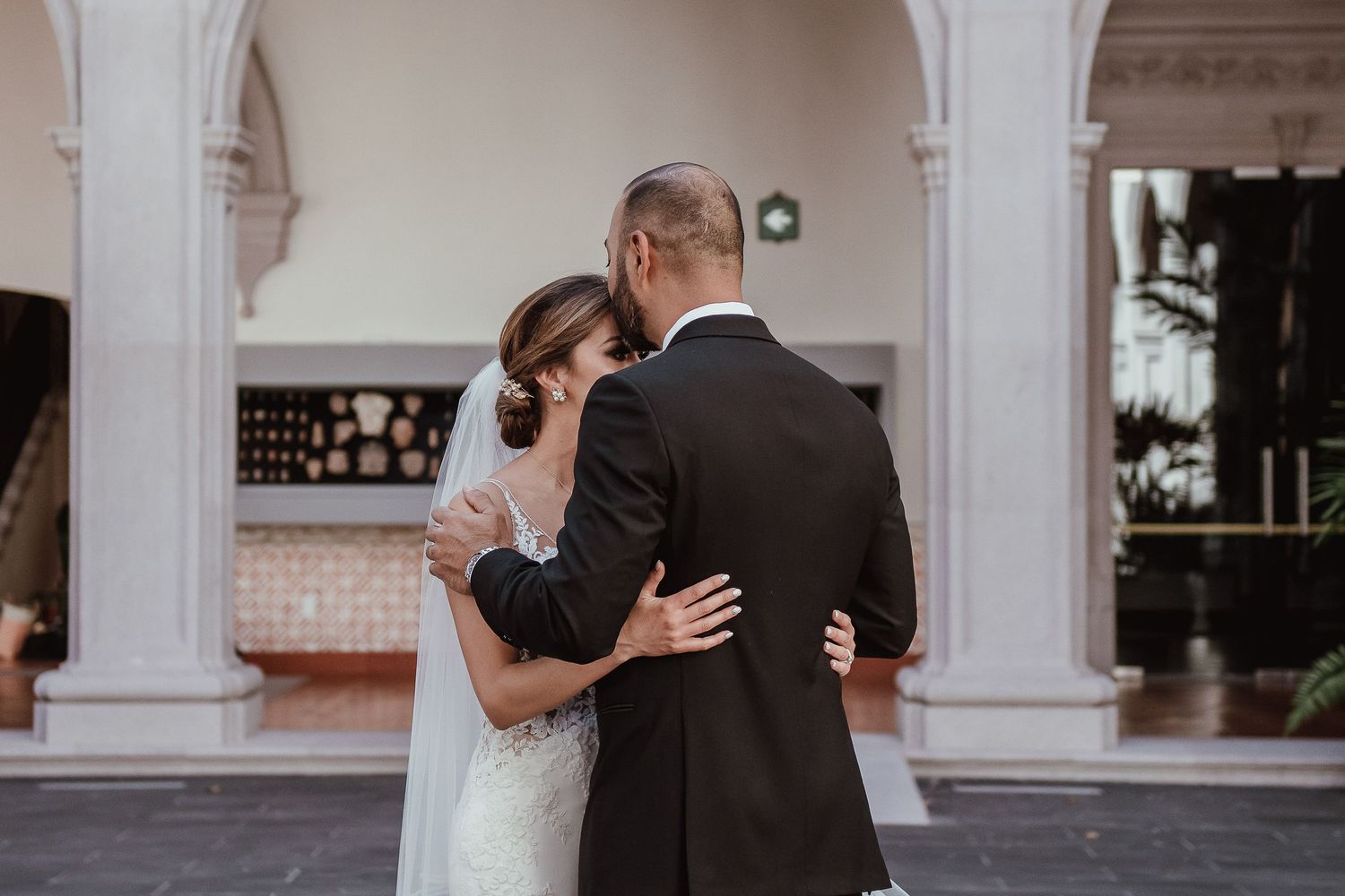 A bride and groom share a tender embrace outdoors at their wedding venue, surrounded by elegant white columns.