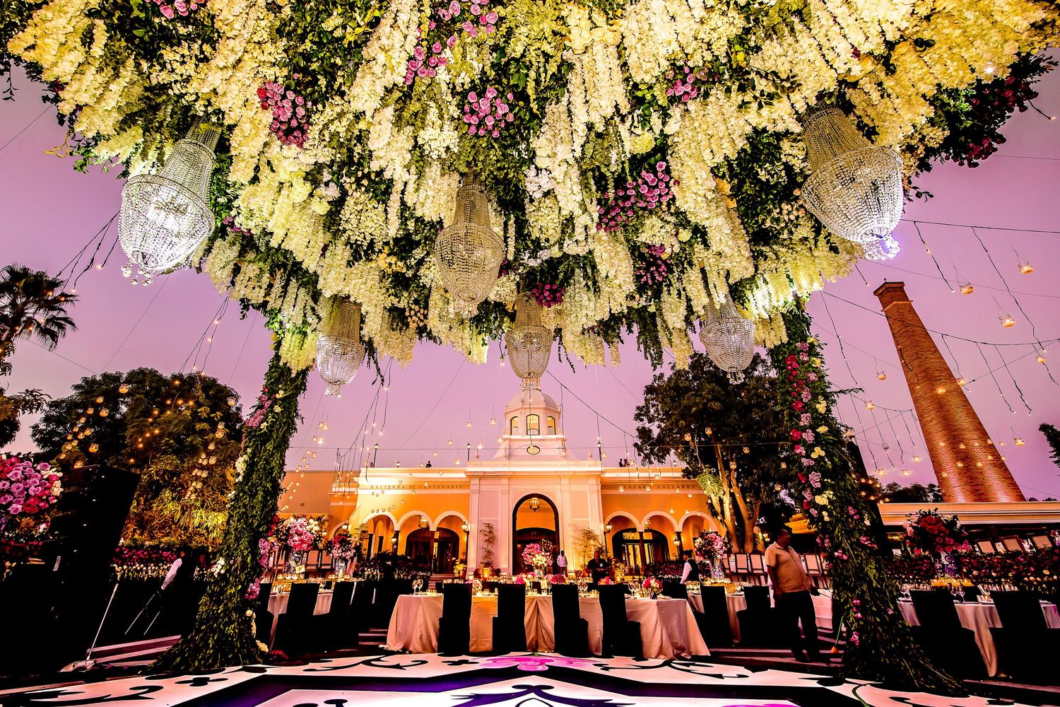 Elegant outdoor wedding reception with lush floral ceiling installation and string lights at dusk.