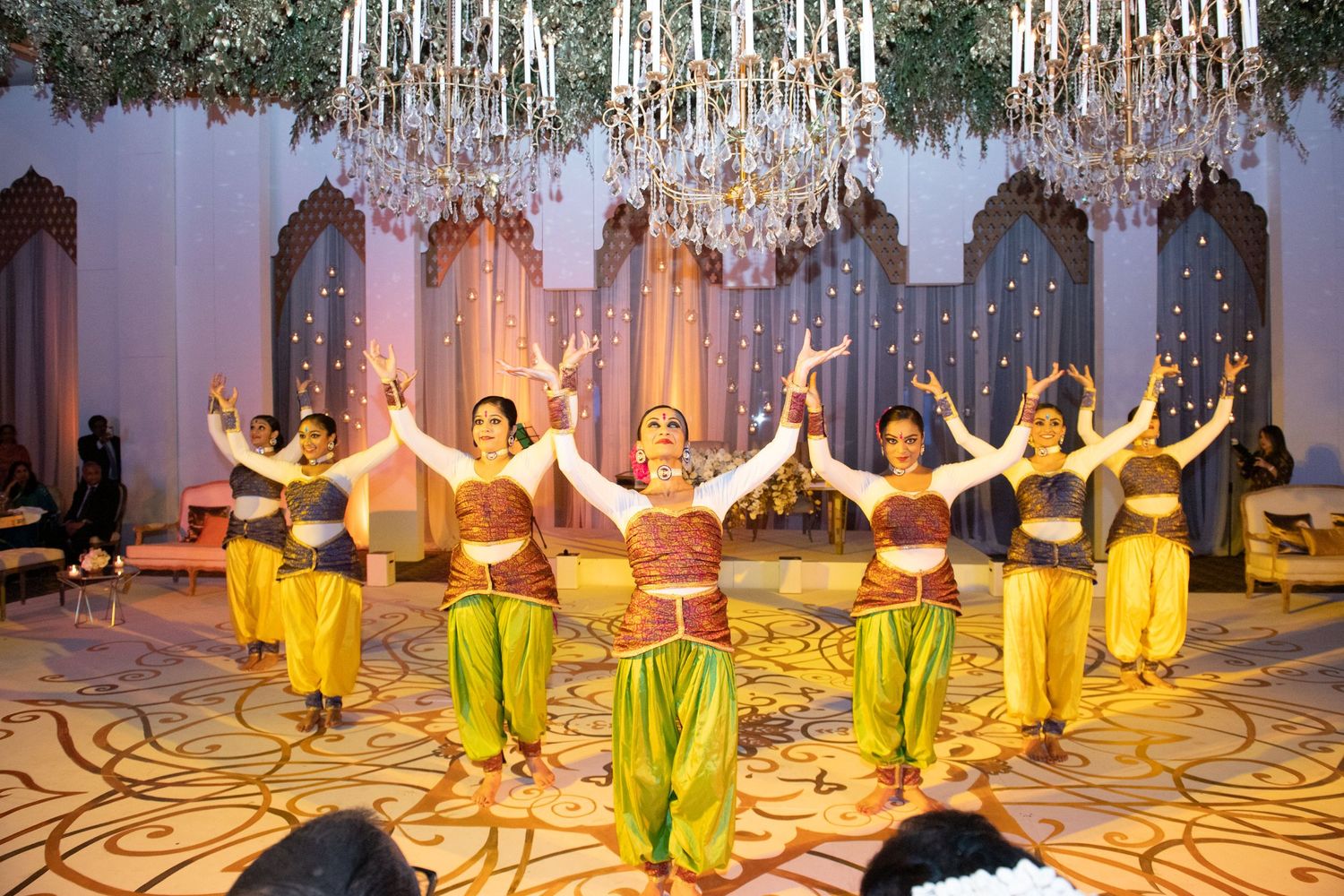 Indian classical dancers in colorful yellow and red costumes perform on a decorated stage at a festive celebration.