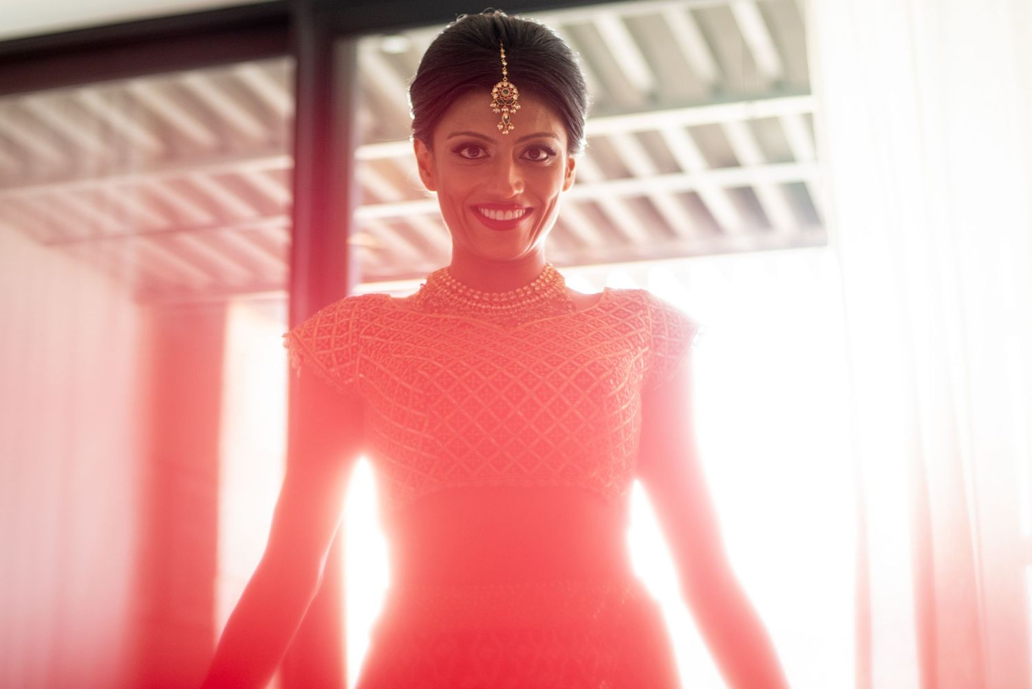 Elegant Indian bride in red beaded gown with maang tikka jewelry smiling in soft natural light.