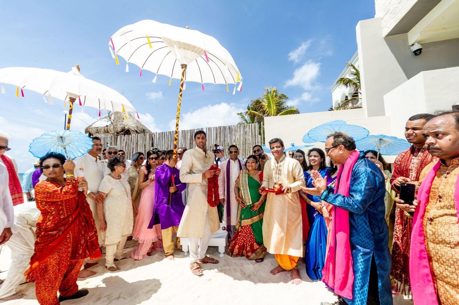 Colorful Indian wedding ceremony on a beach with guests holding white umbrellas under a bright blue sky.
