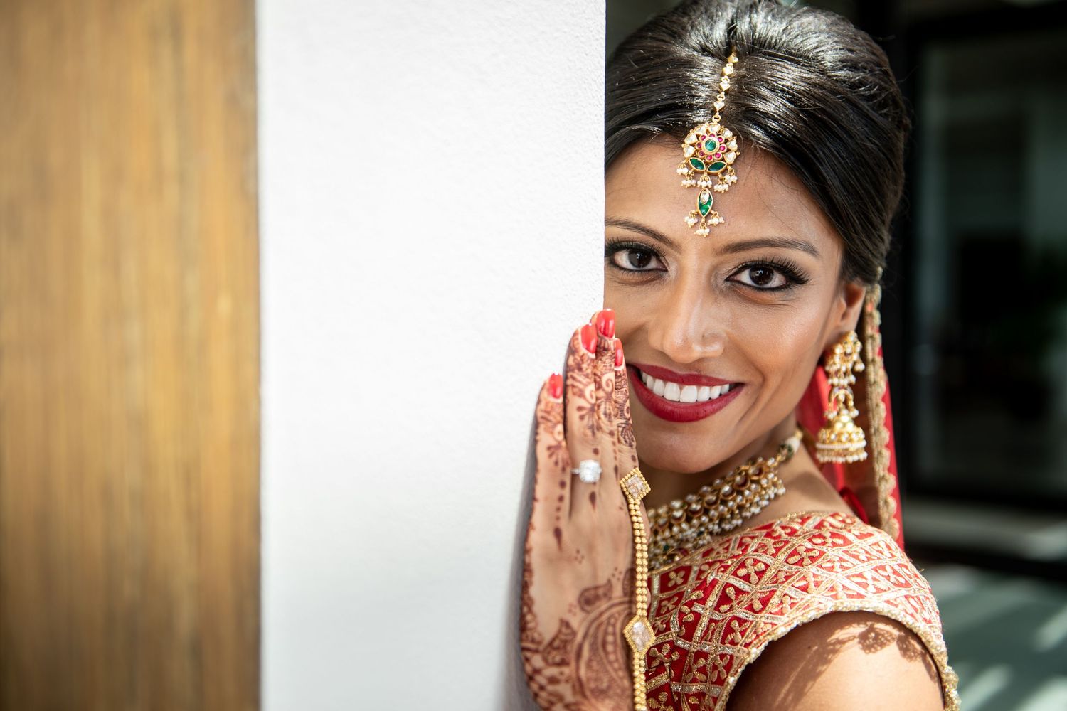 Indian bride in red bridal attire with gold jewelry, mehndi hands, and maang tikka smiling beautifully on her wedding day.