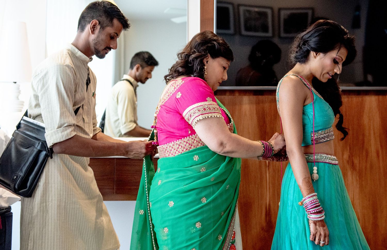 Women in vibrant green and pink Indian saris at a traditional ceremony, with men in traditional attire in background.