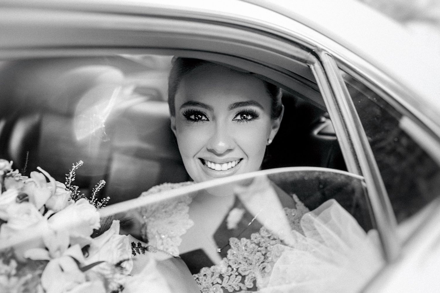 A smiling person in a bridal veil looks out from a car window during a wedding celebration.