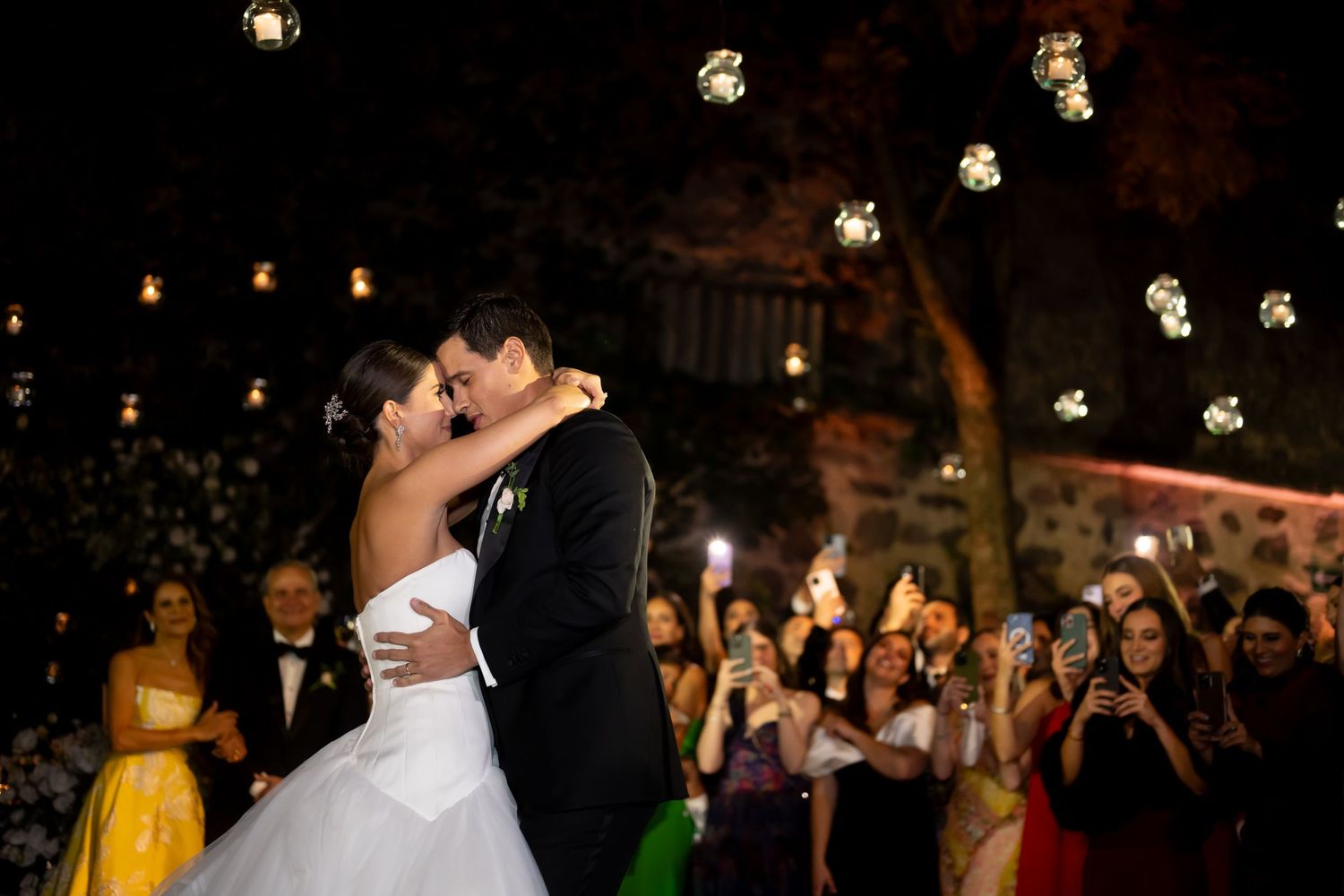 Bride and groom share first dance kiss under glowing string lights while wedding guests celebrate and photograph the moment.