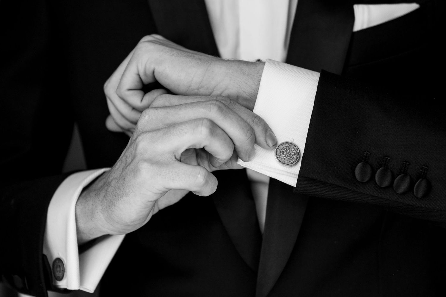 Man in black tuxedo adjusting cufflinks on white dress shirt, black and white photo.
