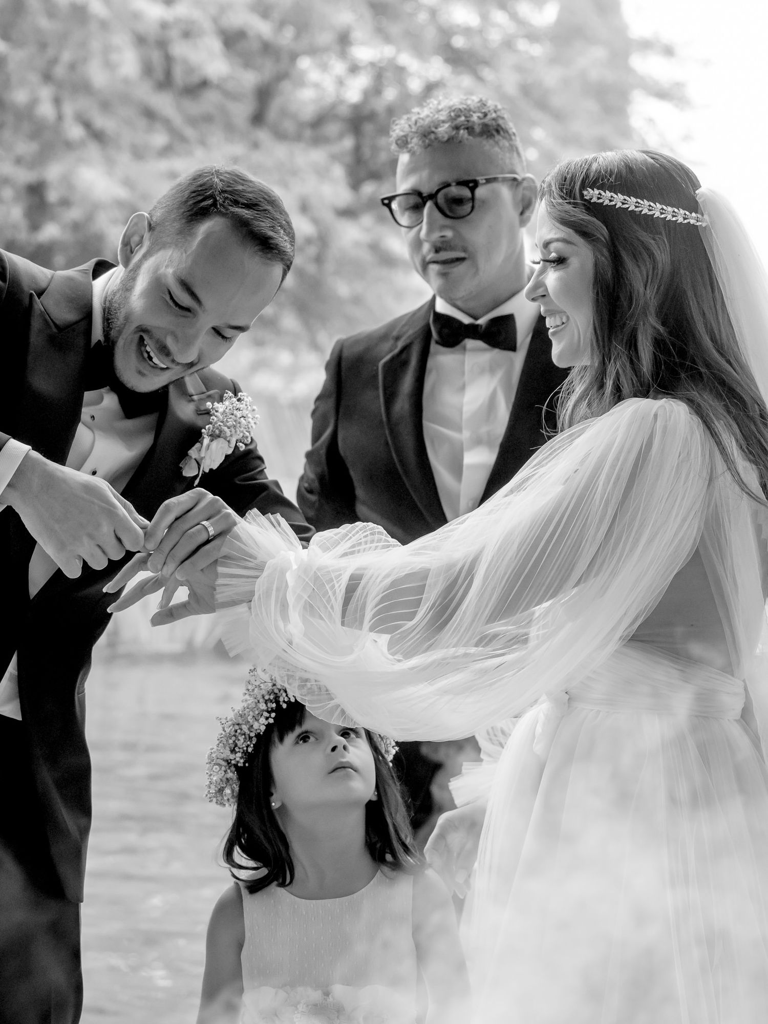 Black and white wedding photo of bride and groom with flower girl and officiant during outdoor ceremony.