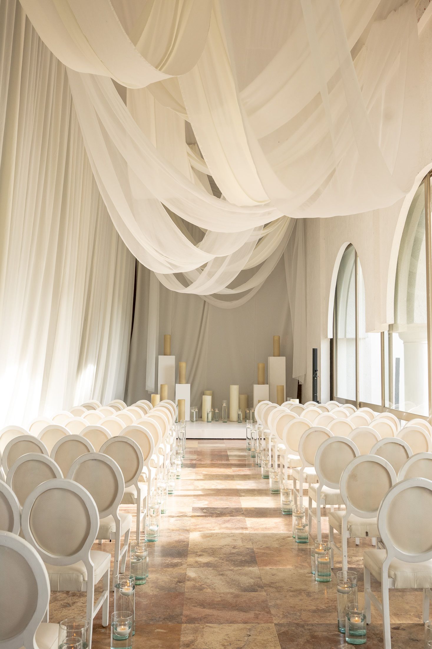 White draped ceremony space with elegant chairs and aisle decorations in a symmetrical layout.