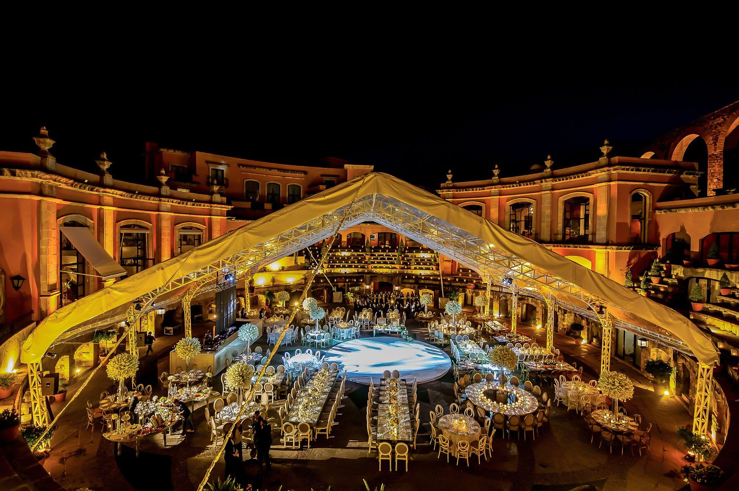 Grand outdoor wedding venue illuminated in golden light with symmetrical table arrangements under architectural canopy.