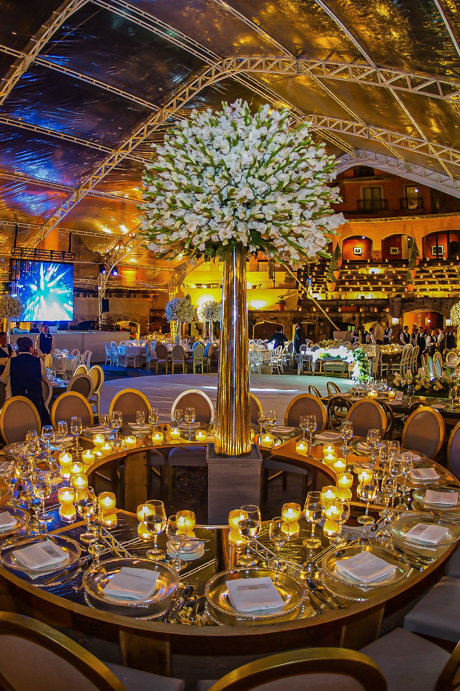 Luxurious wedding table setting with tall white floral centerpiece and golden accents under dramatic lighting.