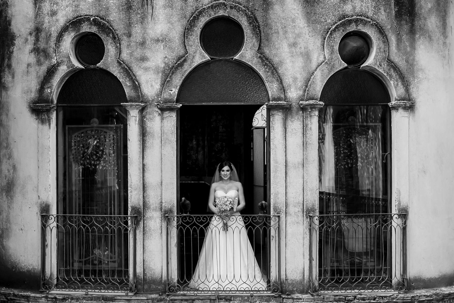 Artistic black and white photo of a bride standing in ornate triple arched doorway with wrought iron gates.