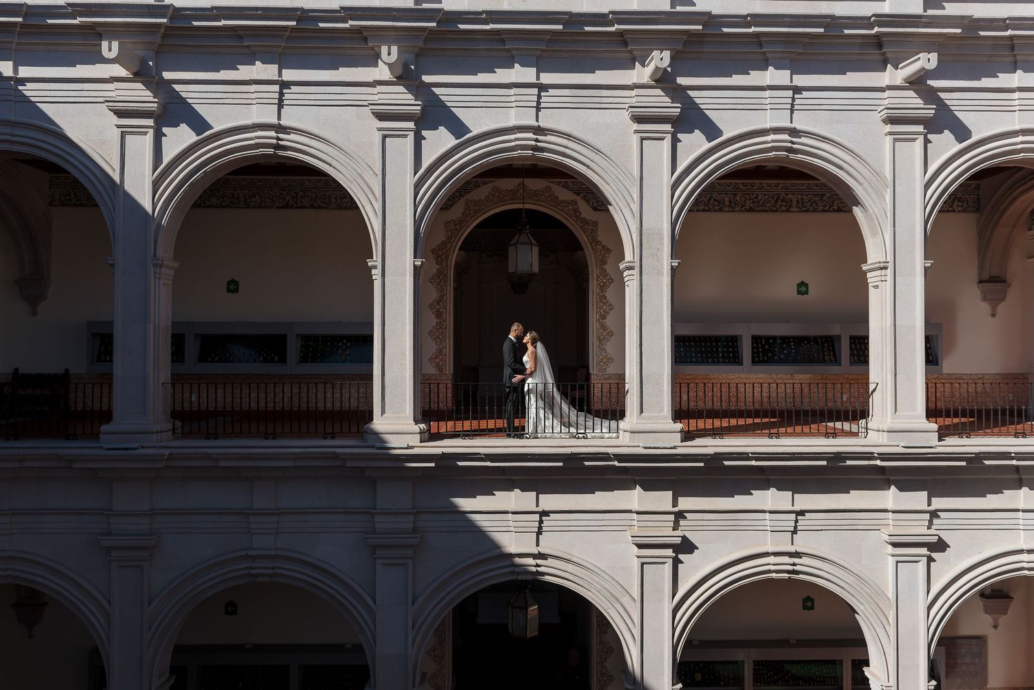 Couple embracing on ornate colonial building balcony with arched arcades in dramatic light and shadow.