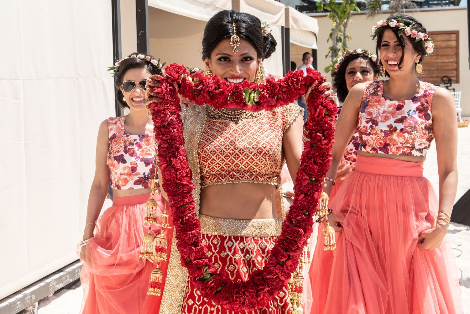 Indian bride in red lehenga holding floral garland with bridesmaids in pink dresses at outdoor wedding ceremony.