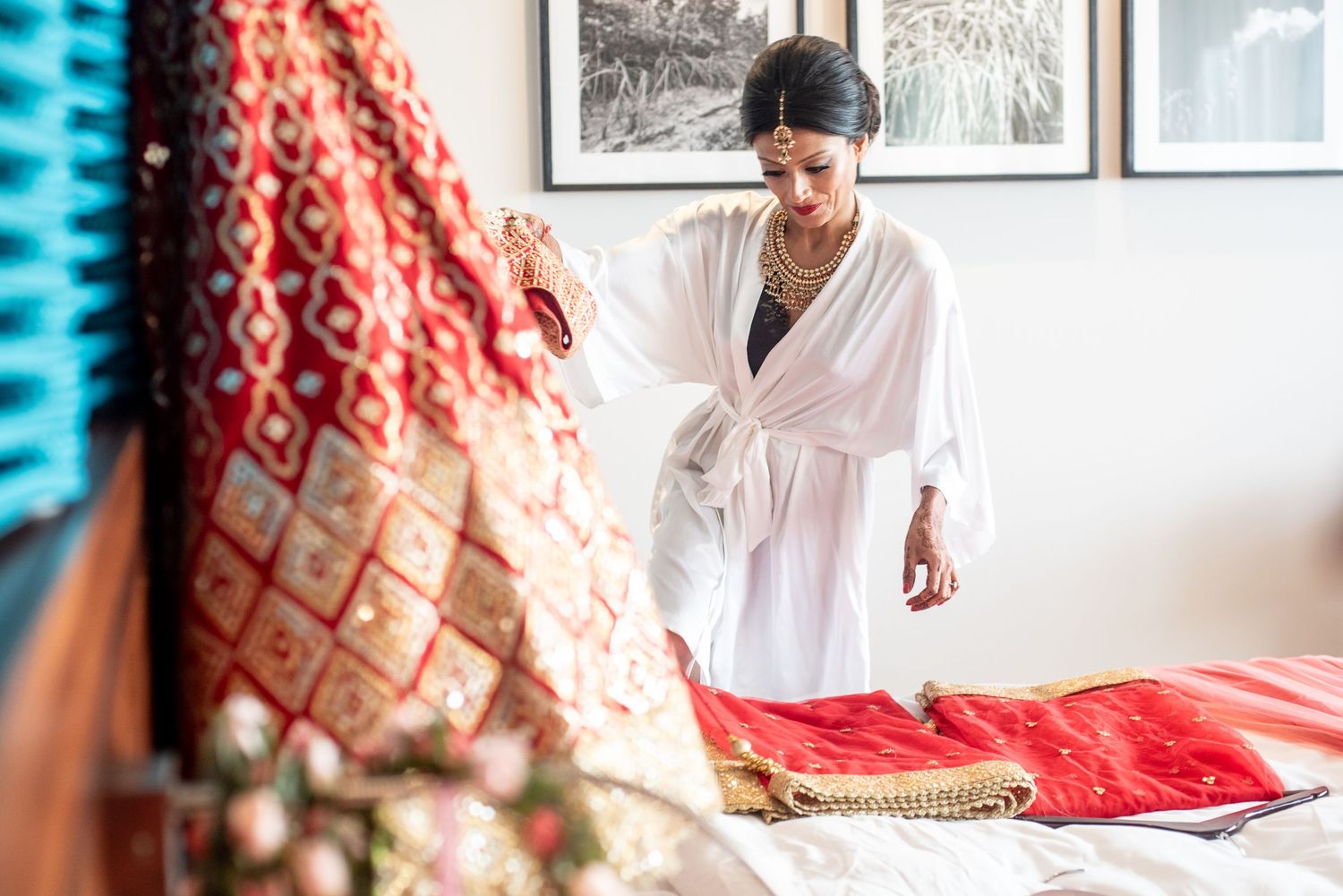 Bride in white robe carefully arranging vibrant red and gold traditional Indian wedding saree on bed.