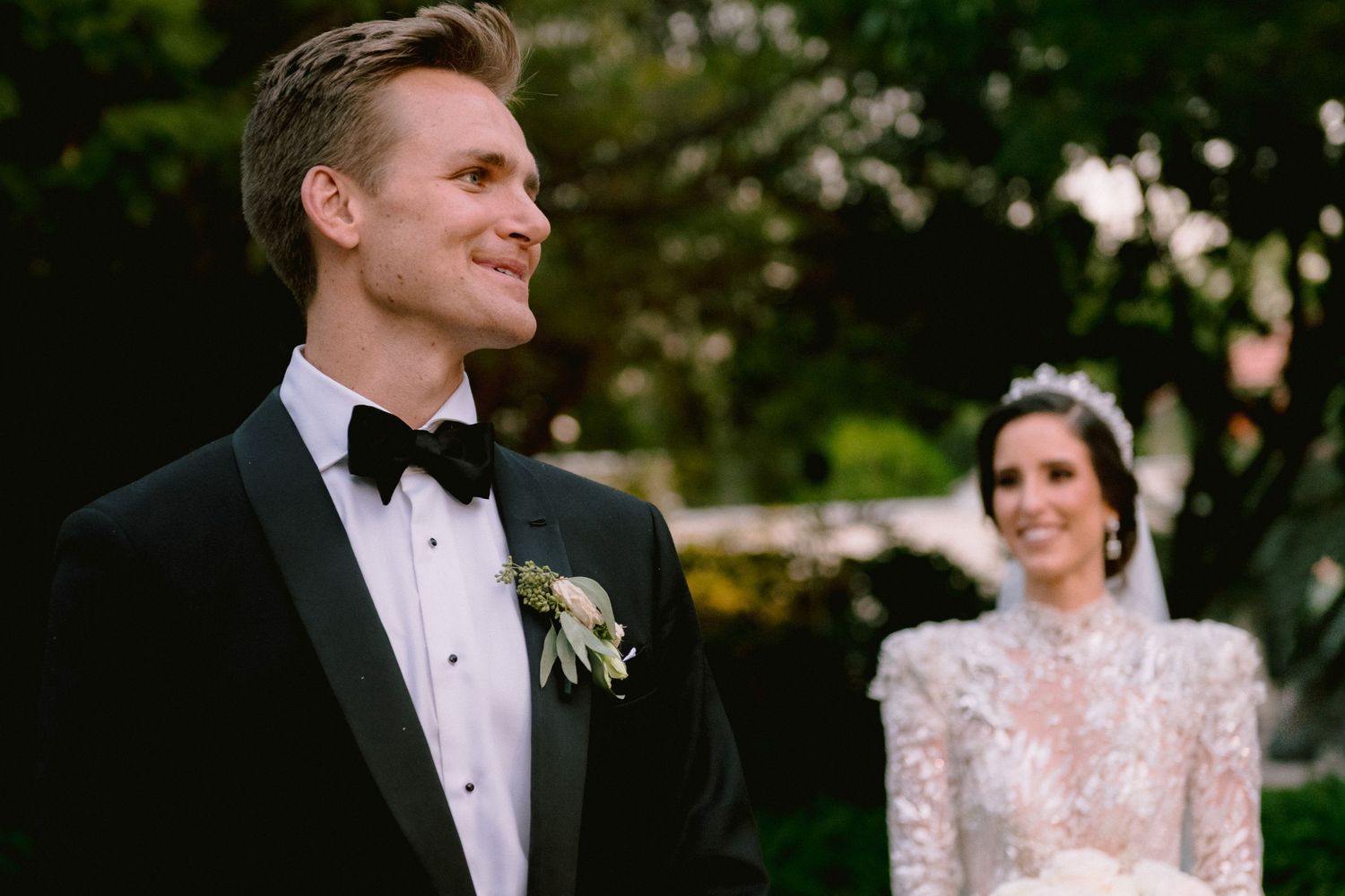 Bride and groom in elegant wedding attire stand outdoors, groom in black tuxedo with boutonniere, bride in lace gown with tiara.