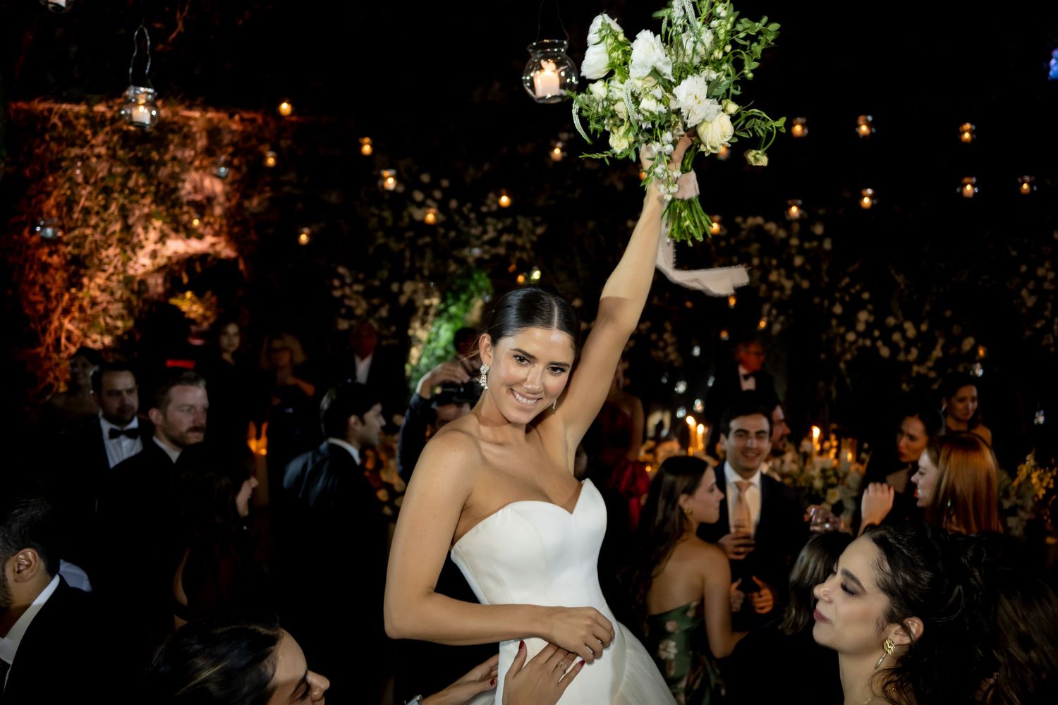 Joyful bride in white strapless gown raises bouquet of white flowers at elegant evening wedding reception.