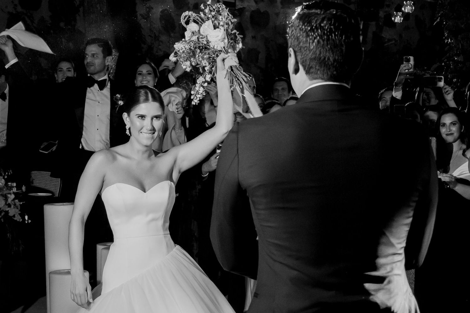 Black and white wedding photo of bride in strapless gown raising bouquet with groom during ceremony.