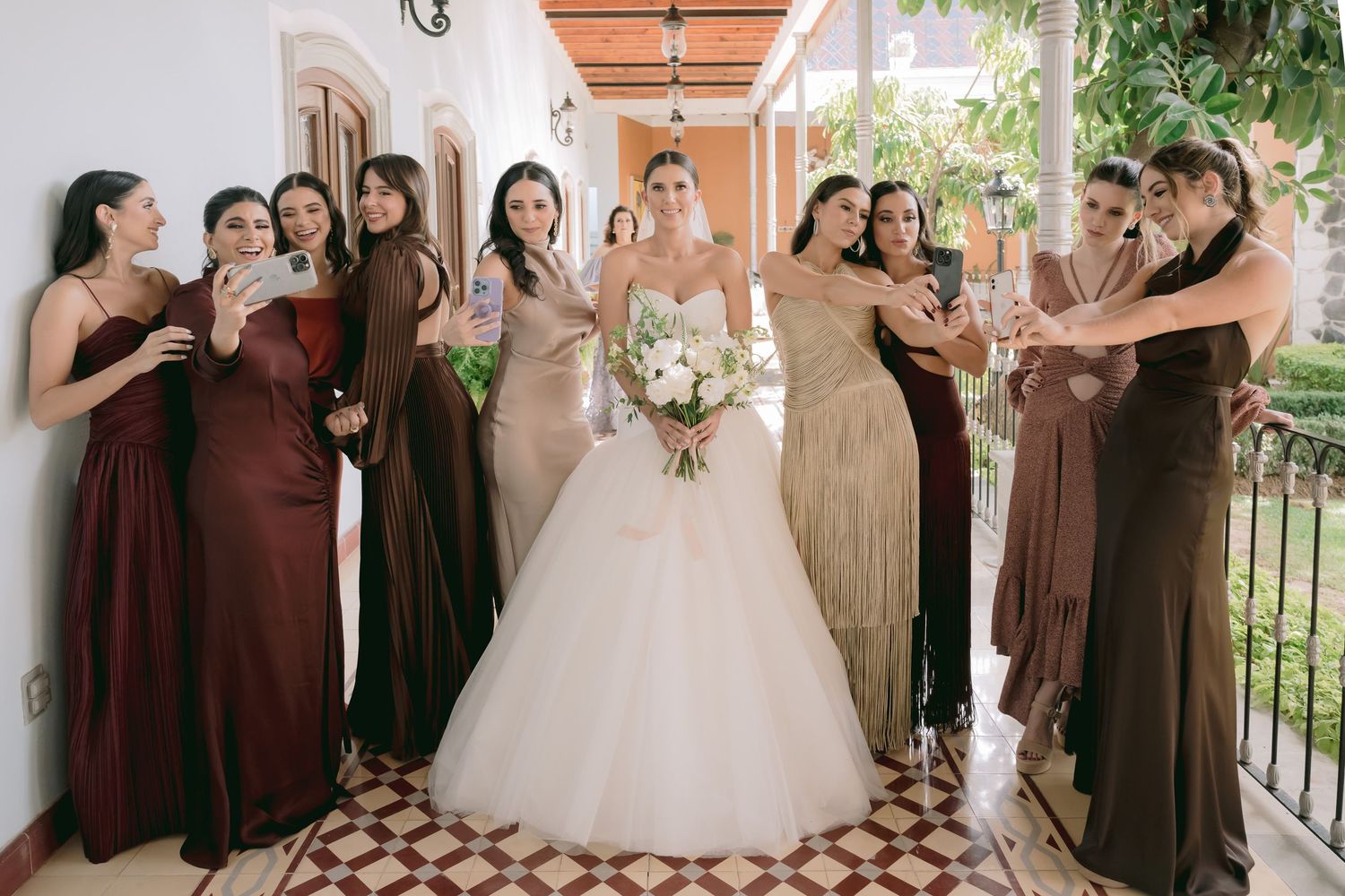 Bride in white ball gown surrounded by bridesmaids in brown and gold dresses on a decorative tiled hallway.