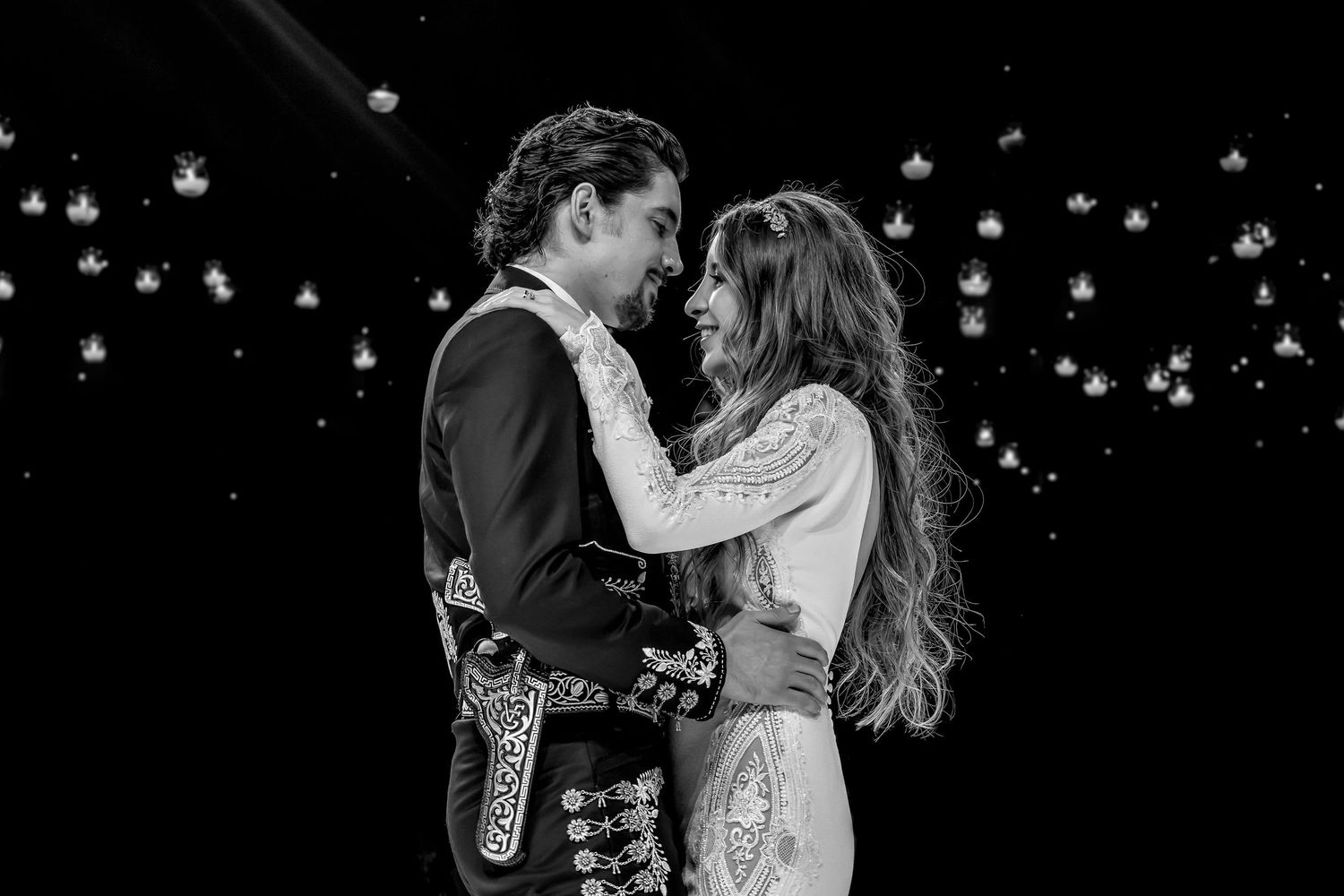 Black and white photo of a couple in formal attire embracing romantically under twinkling lights.