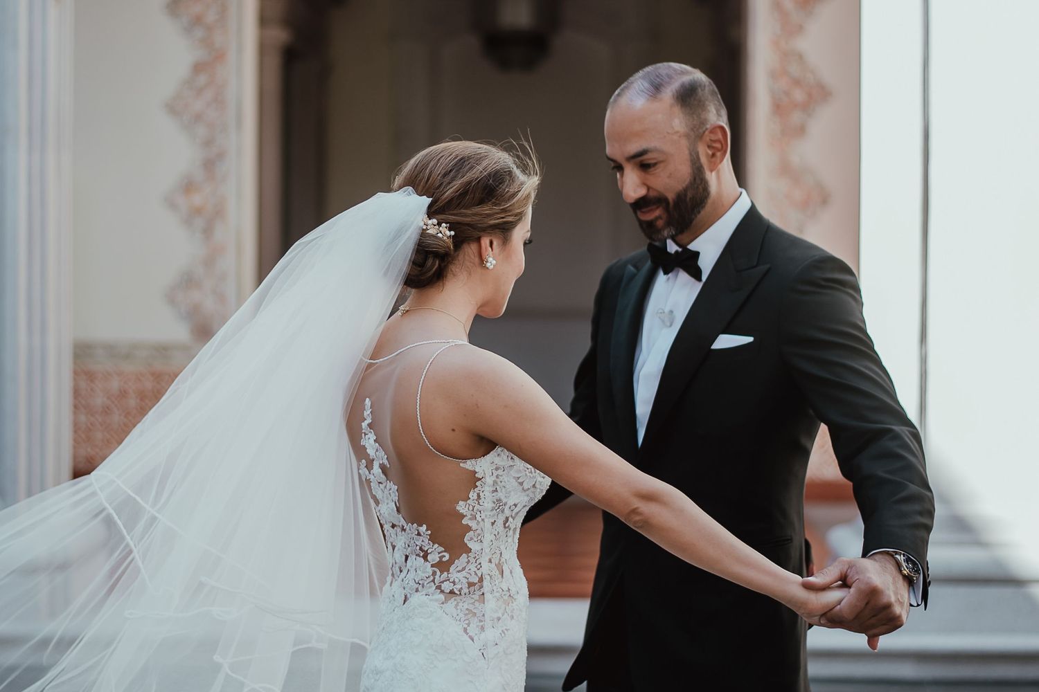 Bride and groom hold hands outdoors, she in lace gown with veil, he in black tuxedo, sharing a romantic moment.