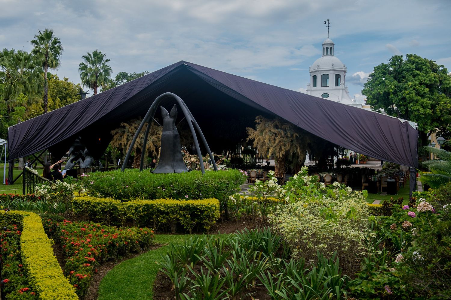 Large black tent structure in a lush tropical garden with manicured hedges and a white building in the background.