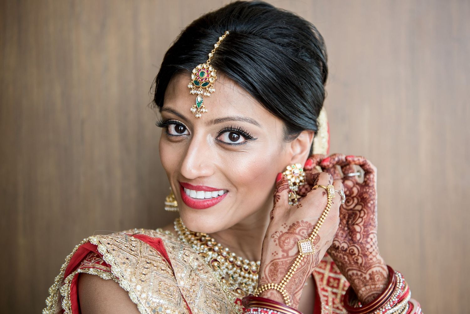 Indian bride in red and gold lehenga with mehndi hands, maang tikka, and necklace smiling elegantly.